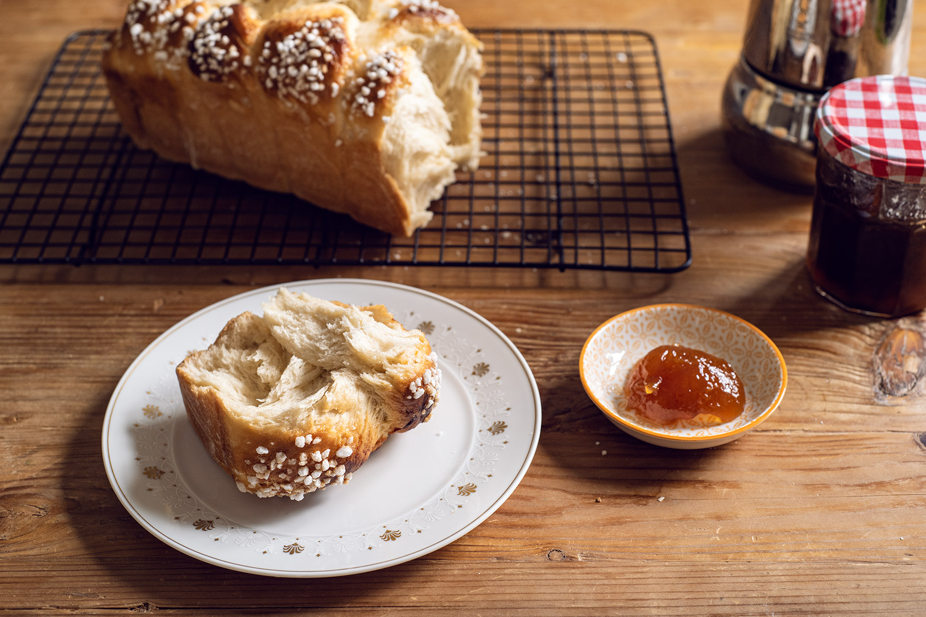 Morceau de brioche dans une assiette, entourée d'un bocal de confiture et d'une cafetière italienne.