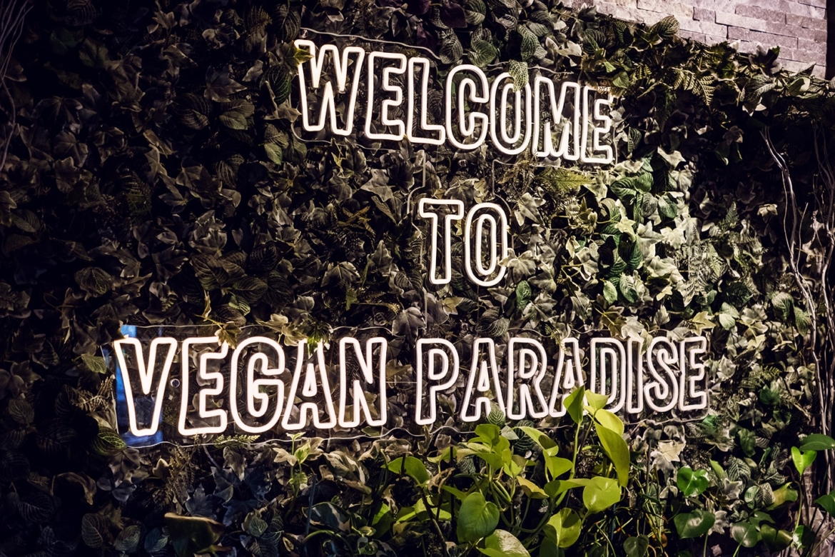 Mur couvert de fausses plantes avec des néons qui composent la phrase "Welcome to vegan paradise".